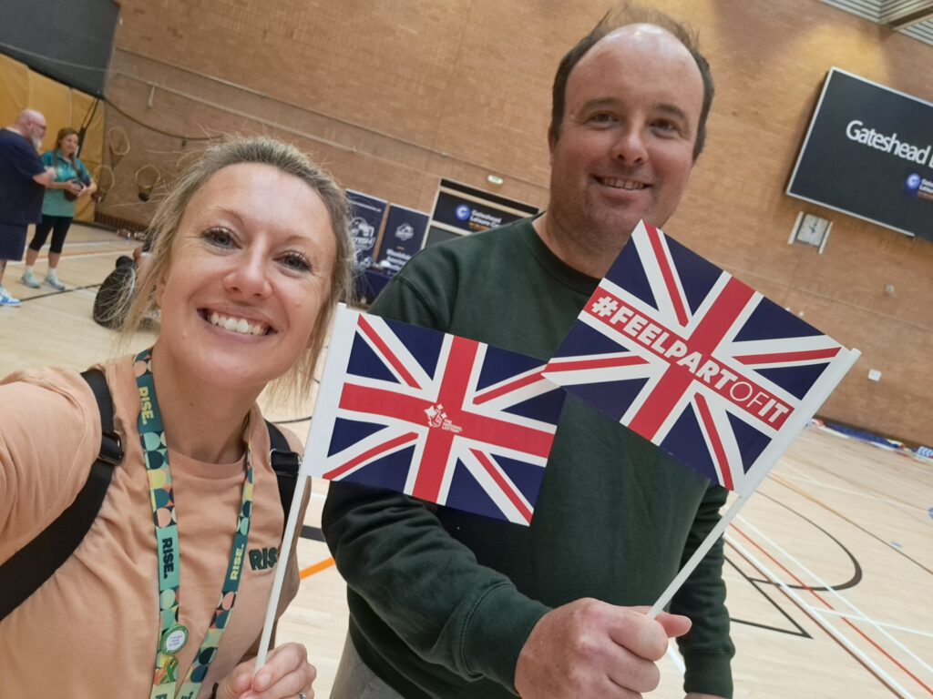 Sarah and Richard at a cerebral palsy sport event, holding small GB flags with #feelpartofit printed on them. 